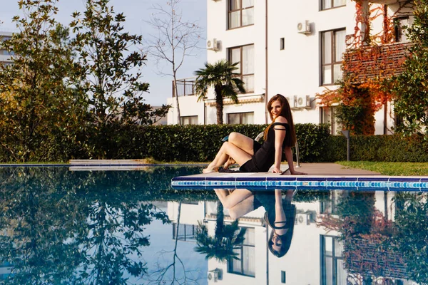 Atractiva chica de pelo largo en vestido negro descansando junto a la piscina azul en su jardín — Foto de Stock