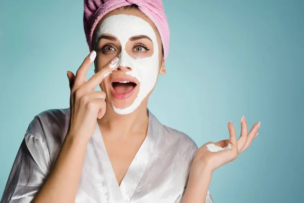 Drôle jeune fille avec une serviette rose sur la tête met sur son visage un masque hydratant blanc — Photo