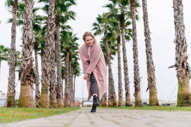 Bahar zevk neşeli kıvırcık saçlı kız pembe bir ceket yanında yeşil palmiye ağaçları, yeşil park boyunca yürür