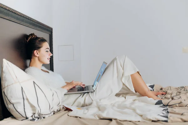 Linda joven en pijama blanco se encuentra en la cama, descansando y ver la película en su computadora portátil — Foto de Stock