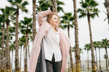 kıvırcık saçlı lüks kadın pembe bir ceket sıcak bahar havanın tadını yeşil park üzerinden yürüyor