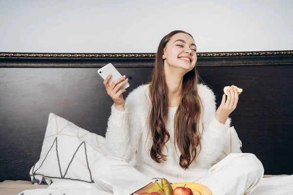 Lachen langhaar meisje zitten in bed, eten van vruchten en smartphone in de hand houden — Stockfoto