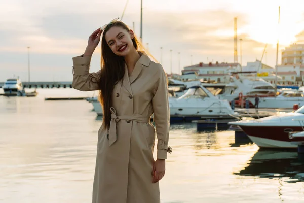 Happy long-haired girl in a beige coat is standing in the seaport and smiling — Stock Photo, Image