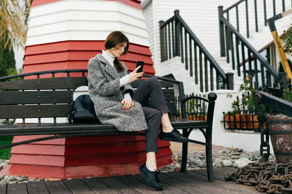 Une fille de la ville aux cheveux foncés en manteau gris s'assoit sur un banc à l'extérieur, regardant dans un smartphone, style de rue — Photo