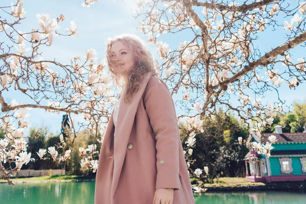 Elegant glamorous woman in a pink coat posing in a park in the sun next to a fragrant magnolia — Stock Photo, Image