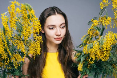 Sarı elbiseli çekici uzun saçlı kız güzel kokulu sarı Mimoza ellerinde tutuyor