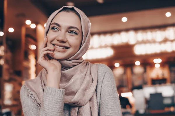 Счастливая арабская девушка с головным платком сидит в уютном восточном кафе, думает о чем-то и смеется — стоковое фото