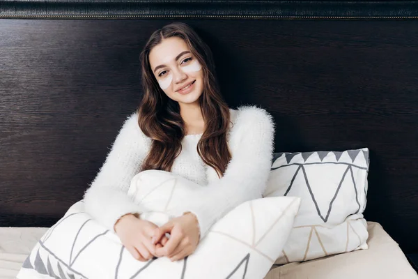 Bela menina de cabelos longos encontra-se na cama no início da manhã, sob os olhos de manchas brancas, sorrindo — Fotografia de Stock