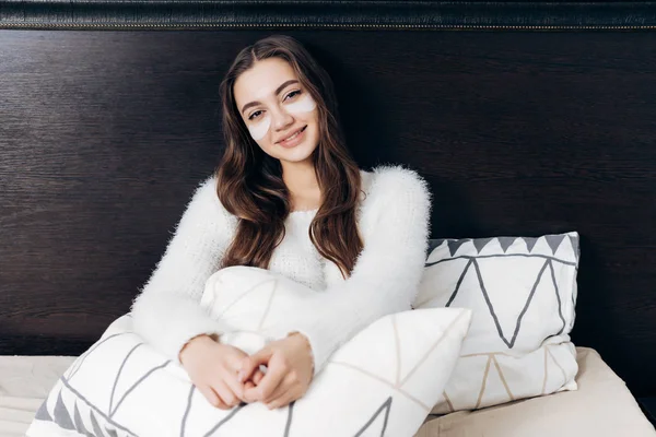 Schöne langhaarige Mädchen liegt frühmorgens im Bett, unter den Augen von weißen Flecken, lächelnd — Stockfoto