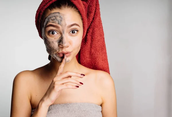 Une jolie jeune fille avec une serviette rouge sur la tête a appliqué un masque d'argile sur la moitié du visage, mis un doigt sur ses lèvres — Photo