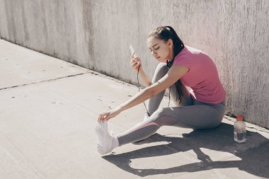 yerde açık havada oturma, kulaklıklar müzik dinlemek ve bacak kasları germe atletik kız