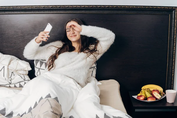 La joven somnolienta yace en la cama. se despertó, sostiene un teléfono inteligente — Foto de Stock