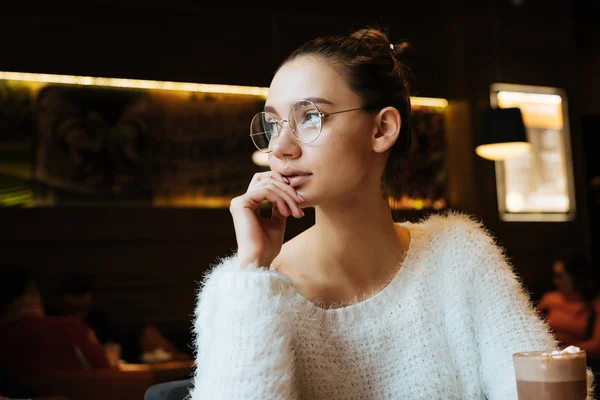 Joven chica pensativa freelancer con gafas sentado en un café y mirando por la ventana, pensando en el trabajo —  Fotos de Stock