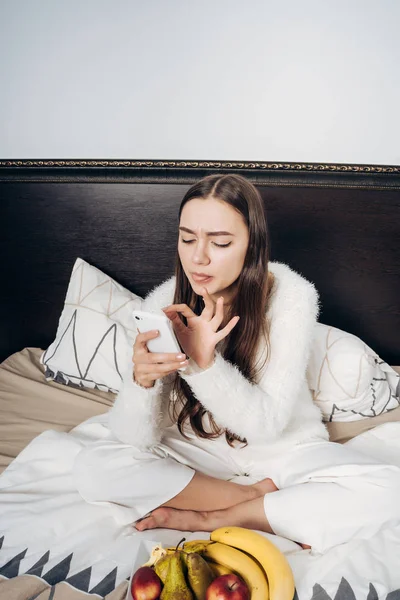 Gadis berambut panjang yang lucu dengan piyama putih duduk di tempat tidur, beristirahat setelah bekerja sehari, menulis pesan di smartphone — Stok Foto
