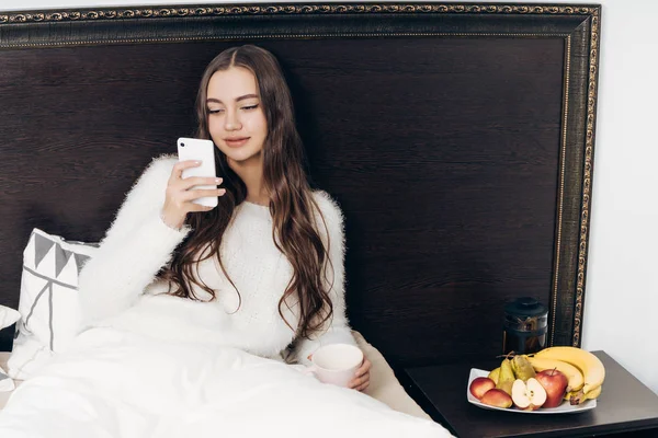 可爱的长头发的女孩坐在床上, 准备睡觉, 看着她的智能手机 — 图库照片