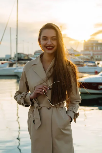 Długowłosy Wspaniała dziewczyna w modny płaszcz posiada okulary w jej ręce, uśmiechając się do portu morskiego — Zdjęcie stockowe