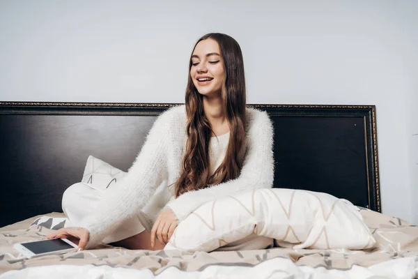 Sonriente chica de pelo largo en pijama se sienta en la cama temprano en la mañana, apaga la alarma en su teléfono inteligente — Foto de Stock