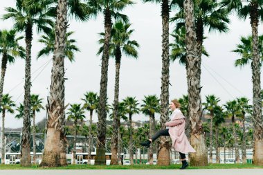 kıvırcık saçlı bir zarif kız pembe bir ceket bahar yeşil park üzerinden yürür ve sıcaklık sahiptir, uzun boylu avuç içi ile birlikte yürür