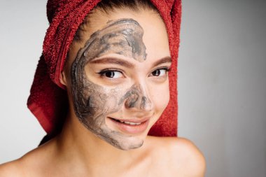 güzel genç kız başında kırmızı bir havlu ile uygulanan bir yararlı kil maskesi yüzünde, gülümseyen