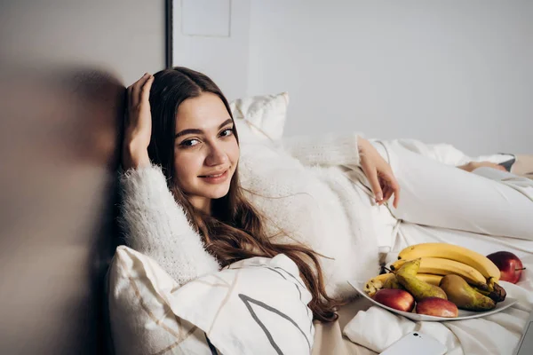 Schönes langhaariges Mädchen liegt im Bett, ruht sich nach einem harten Tag aus und isst Obst — Stockfoto