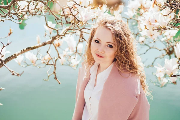 Elegant young girl in a pink coat enjoys blooming fragrant magnolia in the park, by the pond — Stock Photo, Image