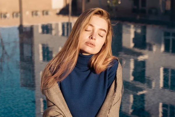 Mooi blond meisje zit bij het blauwe zwembad in de zon, sluit haar ogen en rust — Stockfoto