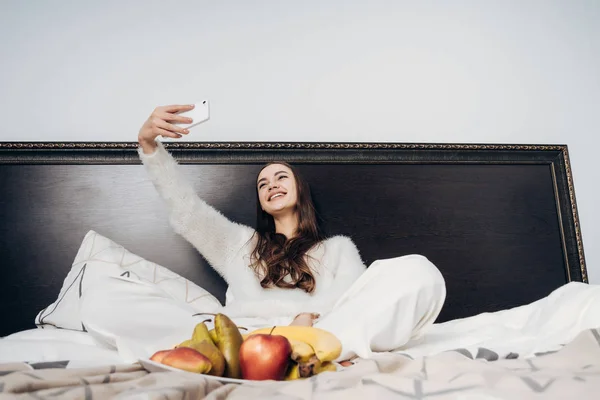 Feliz joven en pijama blanco se sienta en la cama por la mañana, hace selfie en su teléfono inteligente y sonríe — Foto de Stock