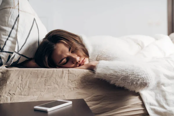 Ein junges Mädchen im weißen Pyjama liegt auf einem Bett, schläft und hört das Klingeln eines Weckers nicht — Stockfoto