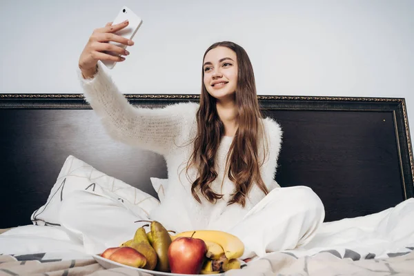 Linda chica de pelo largo en pijama blanco se sienta en la cama temprano en la mañana, hace selfie en su teléfono inteligente — Foto de Stock