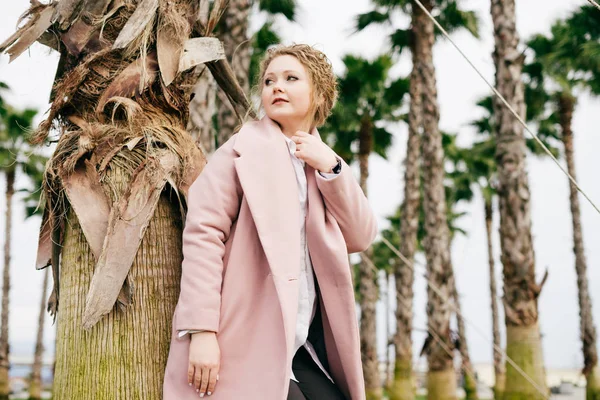 Ricci ragazza elegante in un cappotto rosa passeggiate attraverso il parco verde primaverile e gode il calore — Foto Stock