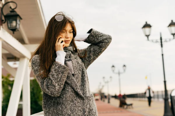 Young dark-haired girl in gray coat walking through the streets of the city, talking on the phone and smiling — Stock Photo, Image