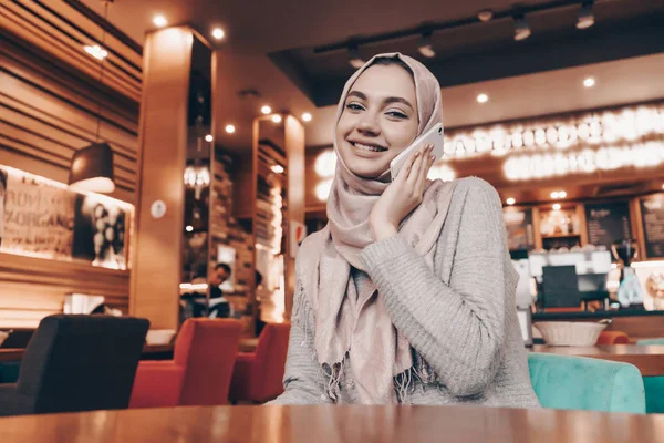 Sorrindo uma garota árabe com um lenço de cabeça sentado em um café oriental, falando ao telefone e sorrindo — Fotografia de Stock