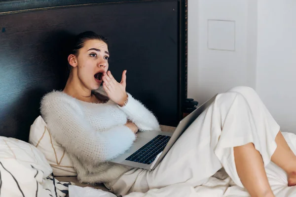 Moe jonge vrouw liggend in bed in witte pyjama, gaapt, werkt in laptop — Stockfoto