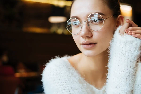 Молодая симпатичная студентка в белом свитере и очках, сидящая в кафе после учебы — стоковое фото