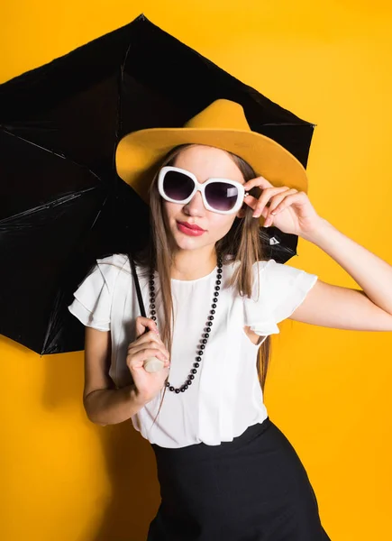 Αυτοπεποίθηση κομψό κορίτσι σε μοντέρνα καπέλο και γυαλιά ηλίου κρατά ομπρέλα και θέτοντας σε κίτρινο φόντο — Φωτογραφία Αρχείου