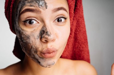 başında kırmızı bir havlu ile güzel bir sürpriz kız yarım yüz için bir spa yordam yararlı kil maskesi uygulanmış