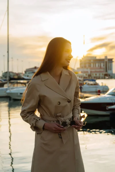 Souriante fille aux cheveux longs dans un manteau beige élégant est debout dans le port de mer dans la luyaah du soleil, va sur un voyage en mer — Photo