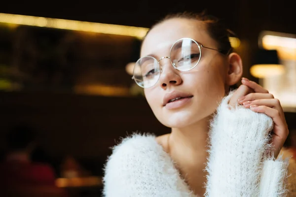 Милий молодий фрілансер дівчина в окулярах і білий светр сидить в кафе і дивиться на камеру — стокове фото