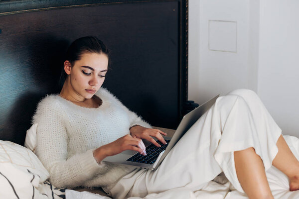 young pensive freelancer girl in white pajamas sitting in bed and working on her laptop in the evening