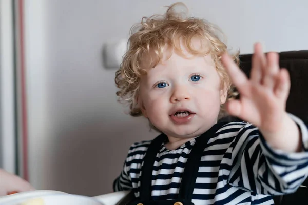 Lindo pequeño niño rizado sentado en una silla alta en la cocina, ojos azules — Foto de Stock