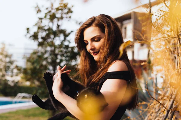 Uma mulher atraente de luxo com cabelos longos segura um gato preto em seus braços, em um jardim nos raios do sol — Fotografia de Stock