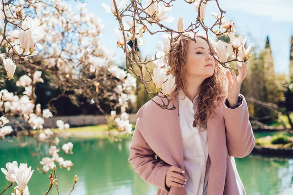 Кучерява молода жінка насолоджується ароматом квітучої магнолії в парку, тепла весняна погода і ставок — стокове фото