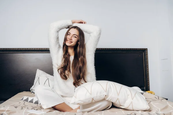 Gelukkig langhaar meisje in witte pyjama zit op het bed, vroeg wakker, strekt zich uit en glimlacht — Stockfoto