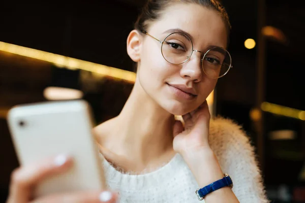 Cute młoda dziewczyna student sobie okulary i biały sweter, siedząc w kawiarni po szkole, trzymając smartfon — Zdjęcie stockowe