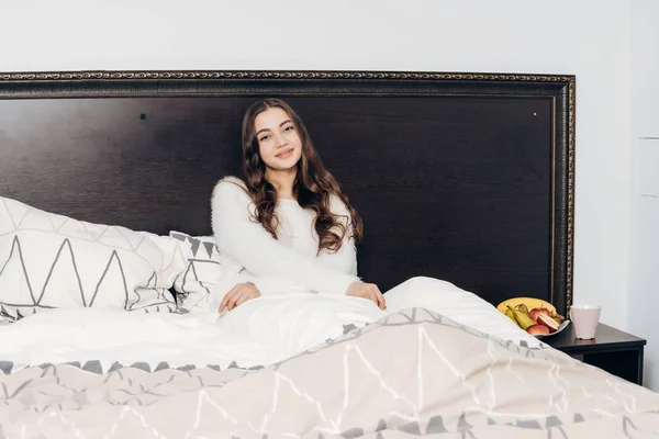 Красивая молодая девушка в белой пижаме сидит на кровати, просыпается рано утром, хорошее настроение — стоковое фото