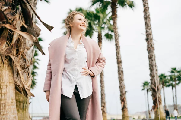 Curly young woman in a pink coat walks through the spring park and enjoys warm weather, laughs — Stock Photo, Image