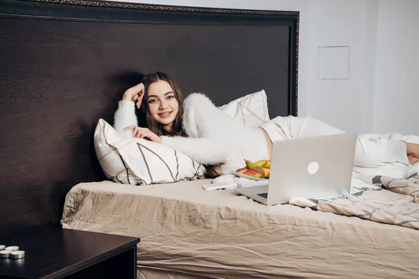 Nettes junges Mädchen im weißen Pyjama liegt frühmorgens im Bett, lächelt und isst Fritten — Stockfoto