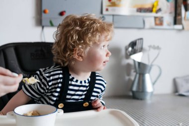 küçük bir kıvırcık çocuk sandalyesi mutfakta oturuyor, annem onu yararlı bir lapa besleniyor