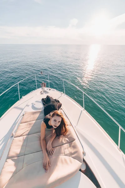 Роскошная девушка в черной стильной одежде лежит на белой яхте и позирует на солнце — стоковое фото