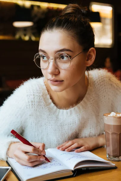 Задумчивая молодая девушка-фрилансер в очках сидит в кафе, работает, пишет в блокноте — стоковое фото